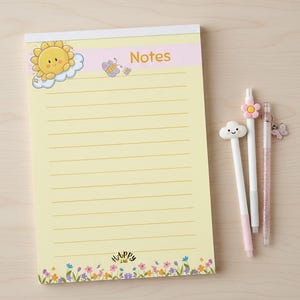 Puede incluir: Un bloc de notas amarillo con páginas rayadas, un sol y una nube, la palabra "Notes" y un borde floral. Tres bolígrafos con adornos decorativos, incluyendo una nube, una flor y un dije de mariposa, están junto al bloc de notas.