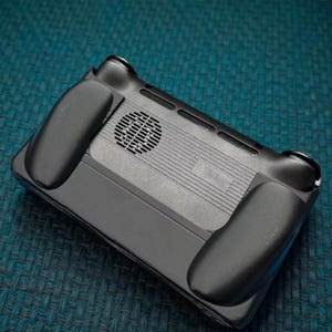 May include: A black handheld gaming console with ergonomic grips. The console features a circular vent and several ventilation slots. The device is designed for comfortable use and enhanced gaming experience.