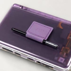 Puede incluir: Una consola de juegos portátil con carcasa morada translúcida y un soporte de tela morado claro para el lápiz óptico. Un lápiz negro con punta plateada está insertado en el soporte. Se aprecian los componentes internos y los puertos del dispositivo.