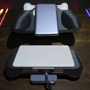 May include: Two handheld gaming console accessories are displayed. The top one has a gray power bank attached. The bottom one has a white tablet attached. Both accessories have black grips with a carbon fiber pattern.