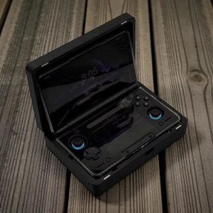 Puede incluir: Dispositivo de juego portátil negro con una pantalla que muestra las 8:05. El dispositivo tiene detalles en azul en los joysticks y botones. El dispositivo está abierto, revelando la pantalla y los controles, sobre un fondo de madera.
