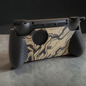 Könnte beinhalten: Ein schwarzer Handheld-Gaming-Konsolengriff mit einer beigefarbenen Rückseite und einem dunkelgrauen Leiterplattenmuster. Der Griff verfügt über texturierte, ergonomische Seitengriffe, eine kreisförmige Belüftungsöffnung und Schultertasten.