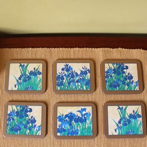 Könnte beinhalten: Sechs quadratische Untersetzer mit einem Blumendesign aus blauen Irisblüten und grünem Laub. Jeder Untersetzer hat einen hellbraunen Rand und einen cremefarbenen Hintergrund. Die Untersetzer sind auf einer strukturierten, beigefarbenen Oberfläche angeordnet.
