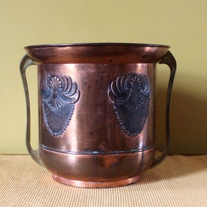 May include: A copper-colored metal container with two handles and decorative embossed designs. The container is cylindrical with a wide opening and sits on a beige placemat. The background is a light green color.