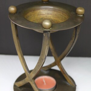 May include: A vintage brass candle holder with a circular base and a bowl-shaped top. The candle holder features four decorative ball accents and curved legs. An orange candle is inside the holder, providing a warm light.