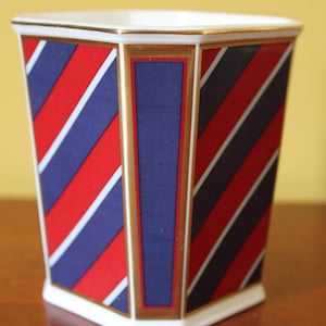 May include: A decorative ceramic planter with a geometric design. The planter features alternating red, navy blue, and white diagonal stripes. Gold trim accents the edges and a navy blue vertical panel. The planter is approximately 5 inches tall.