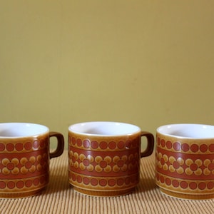 May include: Three ceramic mugs with a brown and orange floral pattern. The mugs have a white interior and a brown handle. The mugs are arranged in a row on a tan surface.