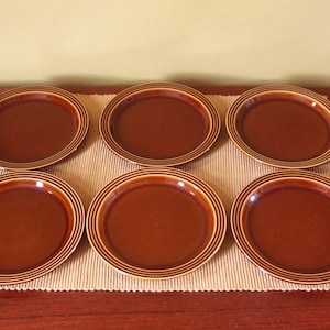 May include: A set of six round ceramic plates in a solid dark brown glaze. Each plate features a series of concentric circular ridges or grooves along the outer rim, creating a textured border around the smooth, flat center of the dinnerware.