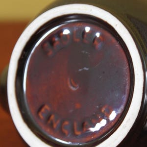 May include: Close-up of the bottom of a dark brown ceramic pitcher with a white rim. The base has a circular, reddish-brown center with the words "Sadler England" embossed in a circular pattern.