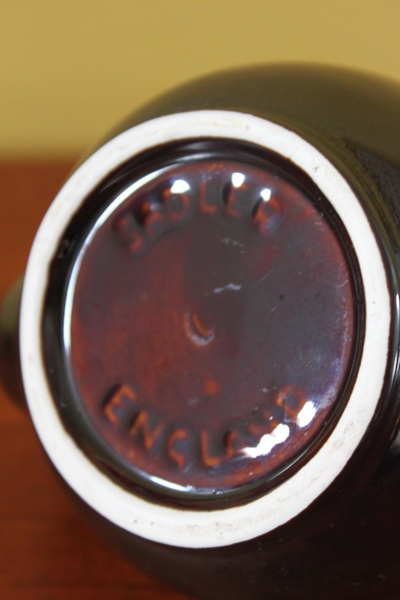 May include: Close-up of the bottom of a dark brown ceramic pitcher with a white rim. The base has a circular, reddish-brown center with the words "Sadler England" embossed in a circular pattern.