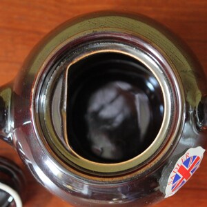 May include: A top-down view of a brown ceramic teapot with a lid. The teapot has a round body, a handle, and a spout. A sticker with the Union Jack flag is visible. The lid is off, revealing the interior.