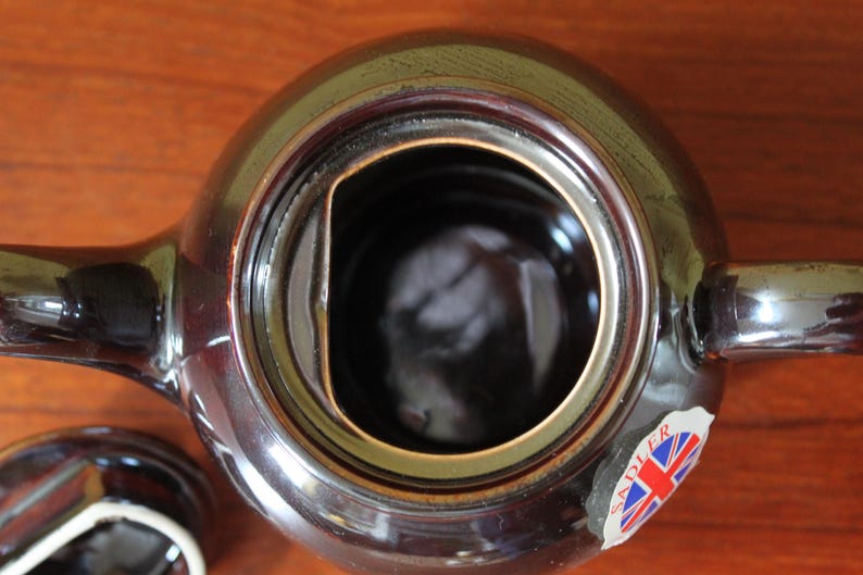 May include: A top-down view of a brown ceramic teapot with a lid. The teapot has a round body, a handle, and a spout. A sticker with the Union Jack flag is visible. The lid is off, revealing the interior.