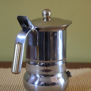 May include: A shiny, stainless steel stovetop espresso maker. The coffee pot has a round base, a cylindrical body, and a lid with a small knob. It features a handle and a hinged lid. The espresso maker is designed for brewing coffee.