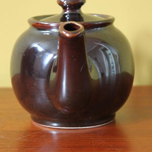 May include: A dark brown ceramic teapot with a matching lid and spout. The teapot has a rounded body and sits on a wooden surface. The teapot is a classic design, suitable for serving tea.