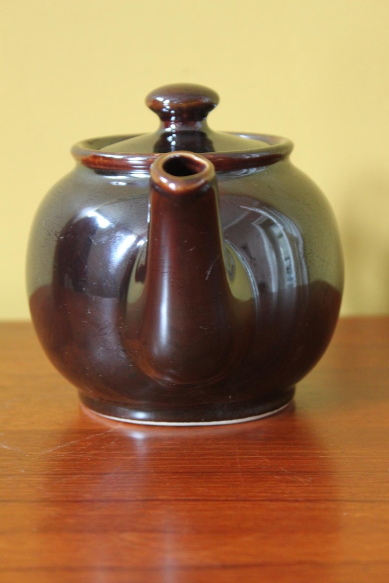May include: A dark brown ceramic teapot with a matching lid and spout. The teapot has a rounded body and sits on a wooden surface. The teapot is a classic design, suitable for serving tea.