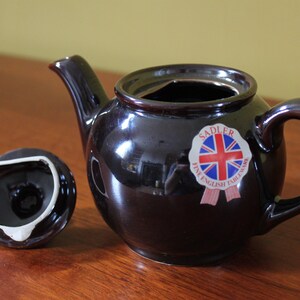 May include: A dark brown ceramic teapot with a matching lid, set on a wooden surface. The teapot has a curved handle and spout. A sticker with a Union Jack flag is on the side. The lid is off to the side.