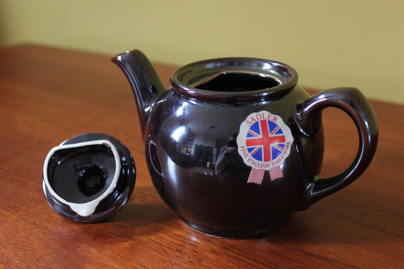 May include: A dark brown ceramic teapot with a matching lid, set on a wooden surface. The teapot has a curved handle and spout. A sticker with a Union Jack flag is on the side. The lid is off to the side.