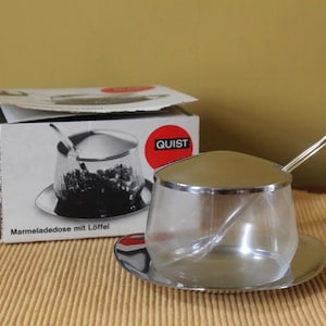 May include: A clear glass jam jar with a polished silver-tone metal lid and matching saucer, accompanied by a silver-tone spoon. The product is displayed next to its original white cardboard box, which features the brand name QUIST and the text Marmeladedose mit Löffel.