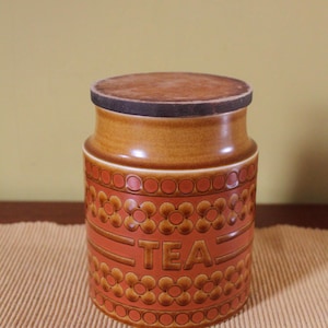 May include: A cylindrical ceramic tea canister featuring a warm orange and brown glaze. The body is decorated with embossed floral patterns and circular motifs, with the word TEA clearly moulded into the centre of the front surface.