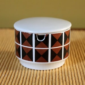 May include: A small, round ceramic container with a lid. The container is white with a geometric pattern of brown and black diamonds. The lid is white, and the container sits on a white base.