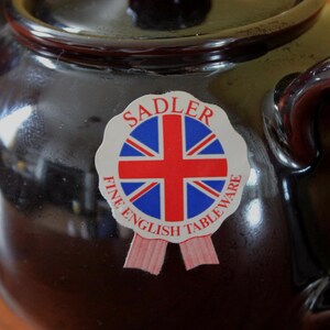 May include: A dark brown ceramic teapot with a lid and handle. A sticker on the front features the Union Jack flag and the words "SADLER FINE ENGLISH TABLEWARE". The teapot has a glossy finish.