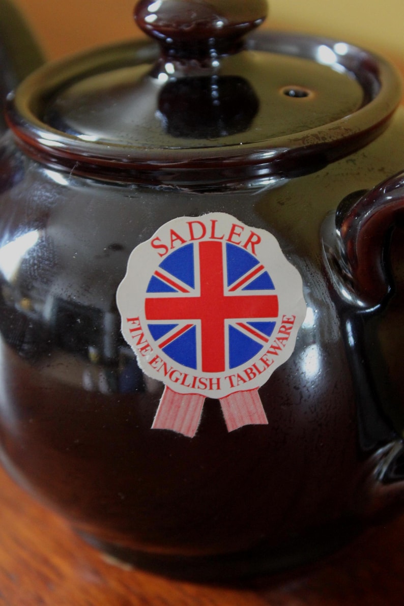May include: A dark brown ceramic teapot with a lid and handle. A sticker on the front features the Union Jack flag and the words "SADLER FINE ENGLISH TABLEWARE". The teapot has a glossy finish.