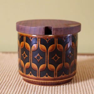 May include: A ceramic jar with a wooden lid. The jar is cylindrical with a brown and black geometric pattern. The lid has a small notch. The jar is likely for storing food items.
