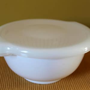 May include: A white, round bowl with a matching lid. The bowl has a smooth, glossy finish and a slightly flared rim. The lid fits snugly over the bowl, creating a tight seal. The bowl is likely used for food storage or serving.