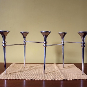 Pode incluir: Um castiçal prateado de cinco braços com um design minimalista. Os castiçais são alargados e ligados por barras horizontais. Está sobre uma esteira clara e texturizada, colocada sobre uma mesa de madeira castanha escura. O fundo é amarelo liso e suave.