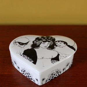 May include: A heart-shaped, white ceramic trinket box with a black and white illustration of three women. The lid features a decorative border and a design of stylized flowers. The box is on a dark wooden surface.