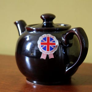 Könnte beinhalten: Dunkelbraune Keramik-Teekanne mit Deckel und Henkel. Die Teekanne hat eine glänzende Oberfläche und einen Aufkleber mit der Union-Jack-Flagge und der Aufschrift "Sadler Fine English Tableware".