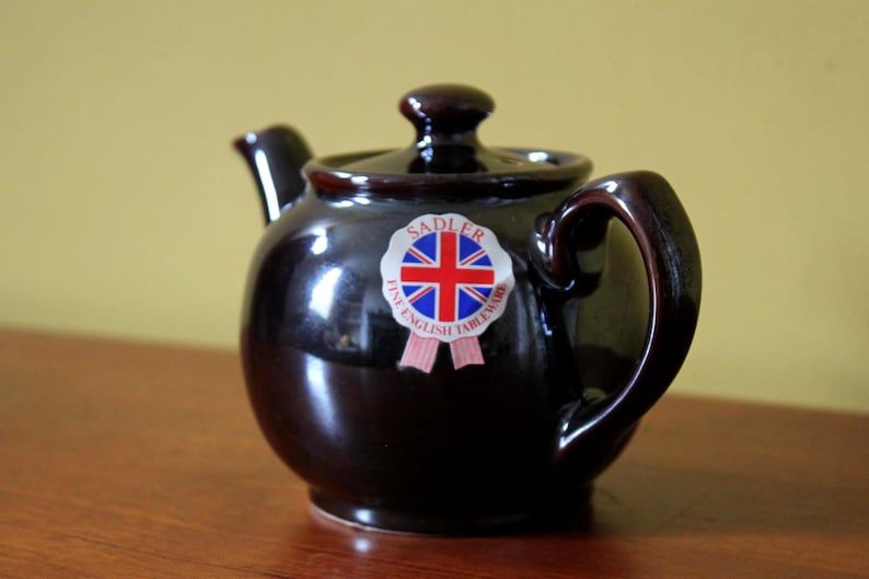 May include: A dark brown ceramic teapot with a lid and handle. The teapot has a glossy finish and a sticker with a Union Jack flag and the words "Sadler Fine English Tableware". The teapot is sitting on a wooden surface.