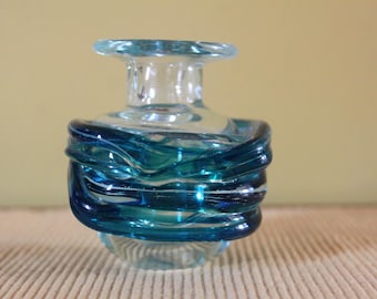 May include: A small clear glass vase featuring a thick, swirling band of deep blue and teal glass wrapped around the centre. The vessel has a wide, flat rim and a rounded base, showcasing a transparent quality with vibrant blue colour accents.