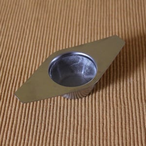 Può includere: Infusore per tè in acciaio inossidabile con manico a forma di diamante e cestello a rete. Progettato per foglie di tè sfuse, viene posto in acqua calda per l'infusione. Sfondo marrone strutturato.