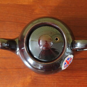 May include: A dark brown ceramic teapot with a lid and handle, viewed from above. The teapot has a spout and a small sticker with a Union Jack flag. The teapot is on a wooden surface.