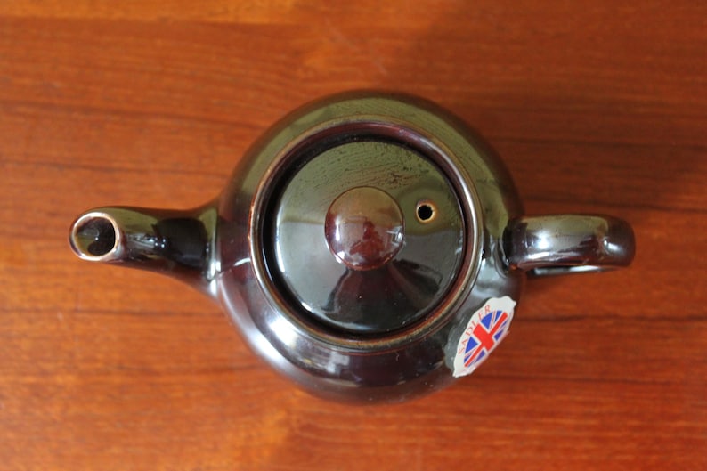 May include: A dark brown ceramic teapot with a lid and handle, viewed from above. The teapot has a spout and a small sticker with a Union Jack flag. The teapot is on a wooden surface.
