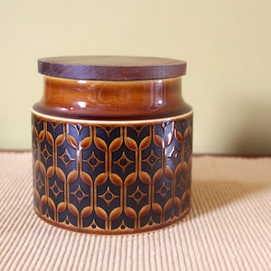 Op de afbeelding: Een cilindrische keramische voorraadpot met een donkerbruin en amberkleurig geometrisch patroon met een herhalend stermotief. De pot is voorzien van een plat, rond, donkerbruin houten deksel dat gelijk ligt met de rand van de container.