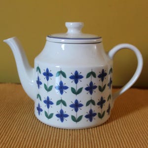 Peut inclure: Une théière en céramique blanche ornée d'un motif répétitif de petites fleurs bleues avec des feuilles vertes. La théière possède un couvercle assorti avec un bord bleu, une anse incurvée et un bec verseur. Le motif est disposé en grille.