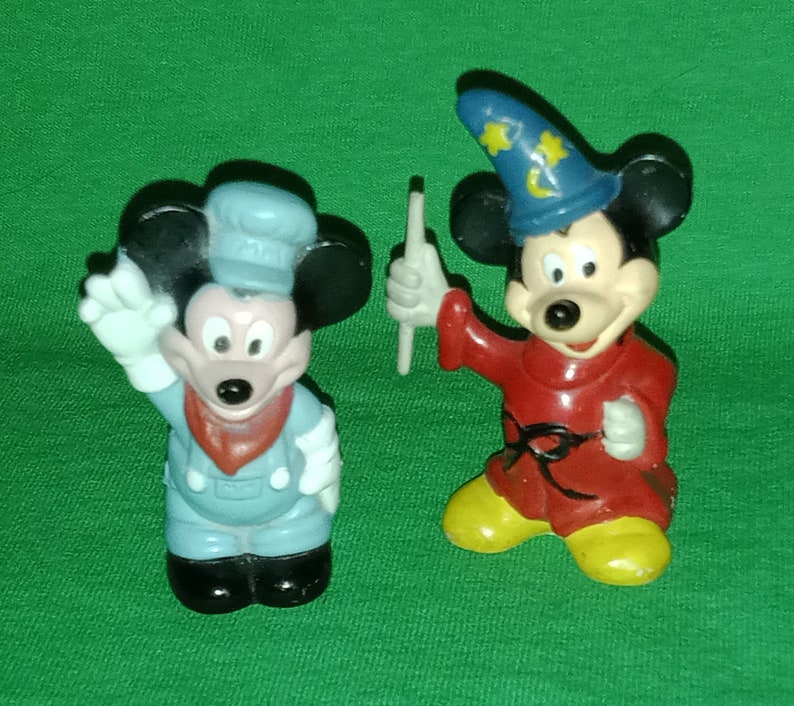 以下が含まれることがあります： ミッキーマウスの小さなプラスチック製フィギュア2体。1体は青い制服と赤いスカーフを着た車掌の姿。もう1体は赤いローブと星がついた青い帽子をかぶったマジシャン姿。