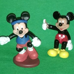 Pode incluir: Duas figuras de pl&aacute;stico do Mickey Mouse. Um Mickey Mouse est&aacute; vestindo um traje esportivo azul e vermelho e o outro Mickey Mouse est&aacute; vestindo shorts vermelhos e sapatos amarelos.