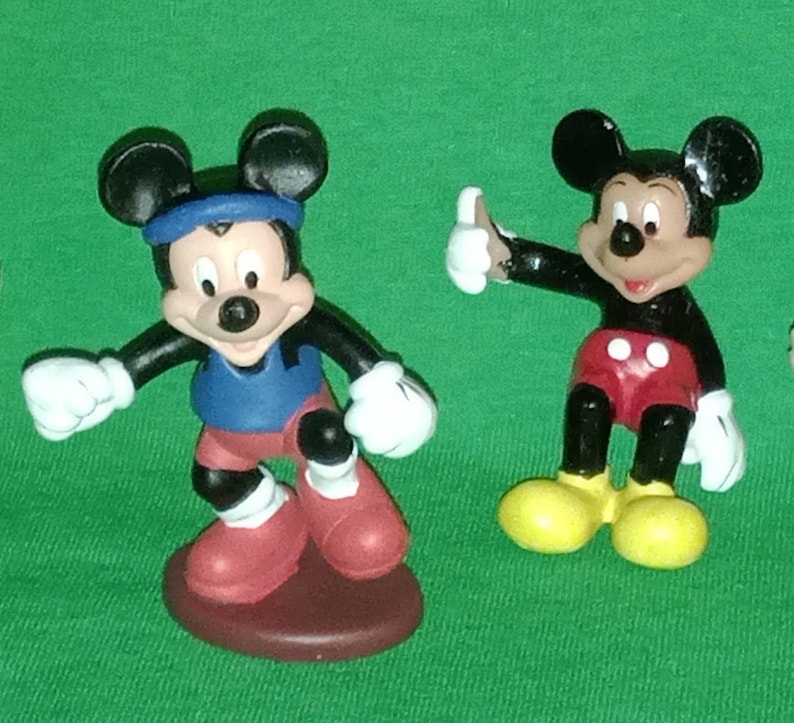 Pode incluir: Duas figuras de pl&aacute;stico do Mickey Mouse. Um Mickey Mouse est&aacute; vestindo um traje esportivo azul e vermelho e o outro Mickey Mouse est&aacute; vestindo shorts vermelhos e sapatos amarelos.