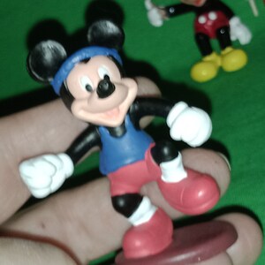Pode incluir: Uma figura de Mickey Mouse preta e vermelha vestindo um traje esportivo azul e branco. A figura est&aacute; em p&eacute; em uma prancha de surf marrom.