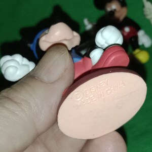Pode incluir: Uma figura de Mickey Mouse vermelha e preta com luvas brancas e uma camisa azul. A figura est&aacute; em p&eacute; sobre uma base rosa com o texto "Disney Made in China".