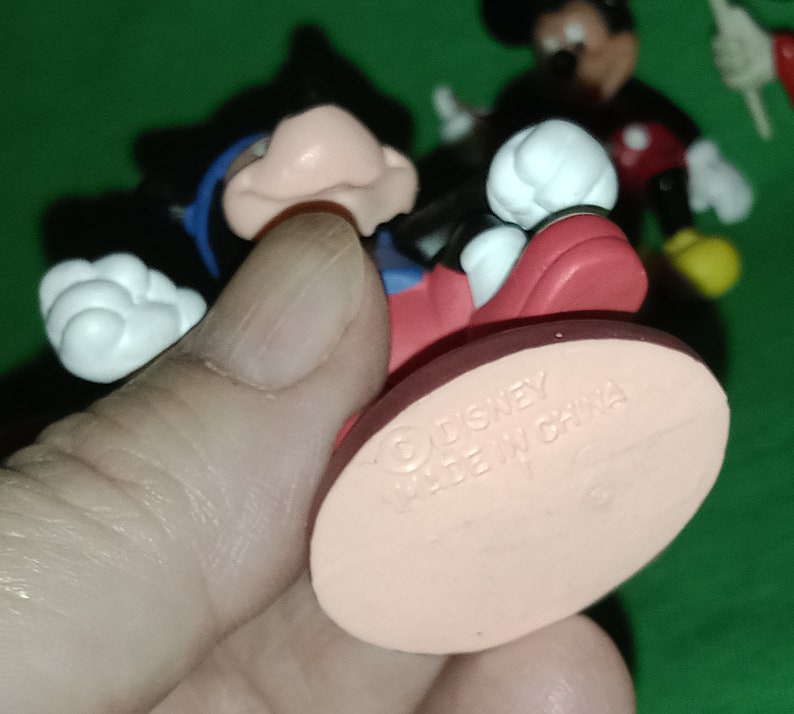 Pode incluir: Uma figura de Mickey Mouse vermelha e preta com luvas brancas e uma camisa azul. A figura est&aacute; em p&eacute; sobre uma base rosa com o texto "Disney Made in China".