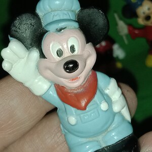 以下が含まれることがあります： 青い帽子とオーバーオールを着たミッキーマウスの青いプラスチック製フィギュア。彼は右手を振っており、首には赤いバンダナをしています。