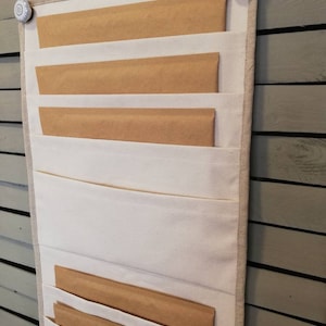 Puede incluir: Un organizador de pared de lona blanca con sobres de papel marrón en cada bolsillo. El organizador está colgado en una pared con pomos decorativos.