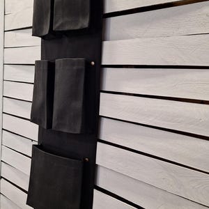 Puede incluir: Un organizador de pared de tela negra con múltiples bolsillos de varios tamaños. El organizador está suspendido de dos pomos decorativos y está montado en una pared de listones blancos. Los bolsillos están diseñados para guardar varios objetos.
