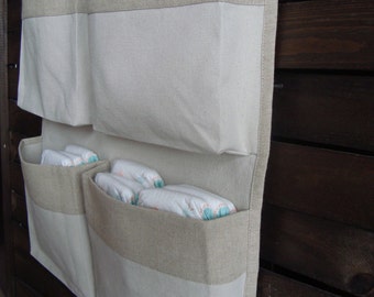 May include: A white canvas hanging organizer with three pockets. The top two pockets have a beige linen trim and the bottom pocket has a beige linen trim. The pockets are filled with towels and diapers.