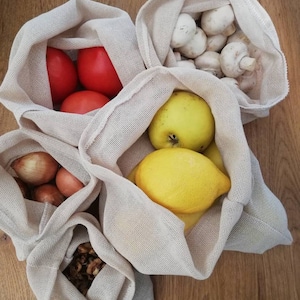 May include: Four white mesh produce bags filled with various fruits and vegetables. The bags contain red tomatoes, white mushrooms, a yellow lemon, a yellow quince, and brown onions.