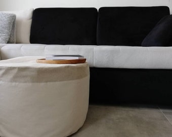 May include: A white and beige round ottoman with a wooden tray on top. The ottoman is in front of a black and white sofa.
